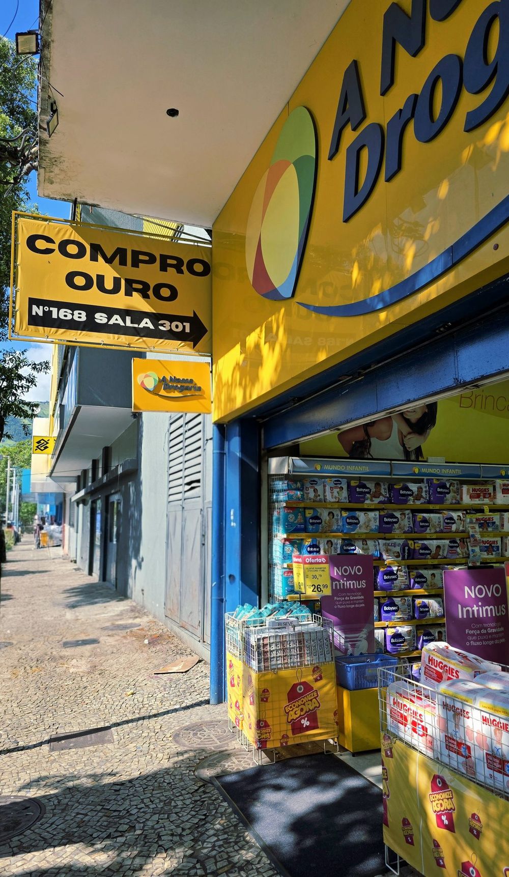 Entrada do prédio com placa Compro Ouro Nº168 Sala 301, ao lado da Nossa Drogaria e do Banco do Brasil